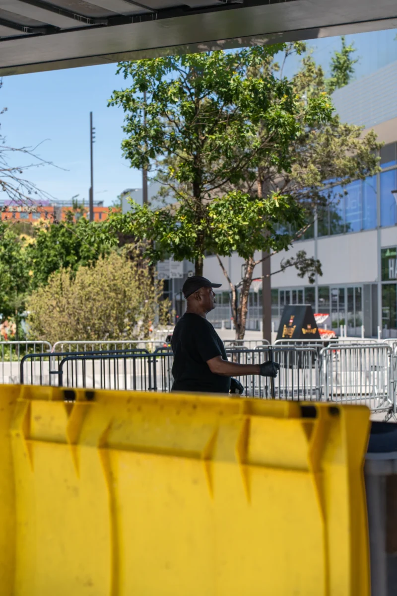 Marc L. · 14/06/2026 · 4672 x 7008 Cette photographie montre une personne assise sur une barrière jaune, en arrière-plan d’un espace urbain. Le sujet principal est cette silhouette, partiellement floue, vêtue de noir.La composition est simple, avec un cadrage serré sur la barrière et un arrière-plan légèrement flou. Les couleurs dominantes sont le jaune vif de la barrière et les tons naturels des arbres et bâtiments en arrière-plan. La lumière semble naturelle, probablement en journée, avec des ombres douces.L’ambiance est calme et un peu mélancolique, évoquant une pause ou une réflexion dans un cadre urbain banal. Le style est réaliste et dépouillé.