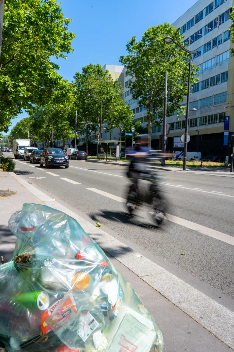 Marc L. · 14/06/2026 · 4672 x 7008 Cette photographie met en scène un sac plastique transparent rempli de déchets (verres, bouteilles) posé sur un trottoir, en premier plan. En arrière-plan, un motard flou traverse une rue urbaine bordée de bâtiments modernes et d’arbres.Le cadrage est serré sur les déchets, contrastant avec l’arrière-plan flou. La lumière est naturelle et vive, suggérant un jour ensoleillé. Les couleurs dominantes sont les verts des arbres, les gris des bâtiments et les teintes claires des déchets transparents. Le style est documentaire et réaliste, évoquant une ambiance urbaine moderne et une réflexion sur la pollution.