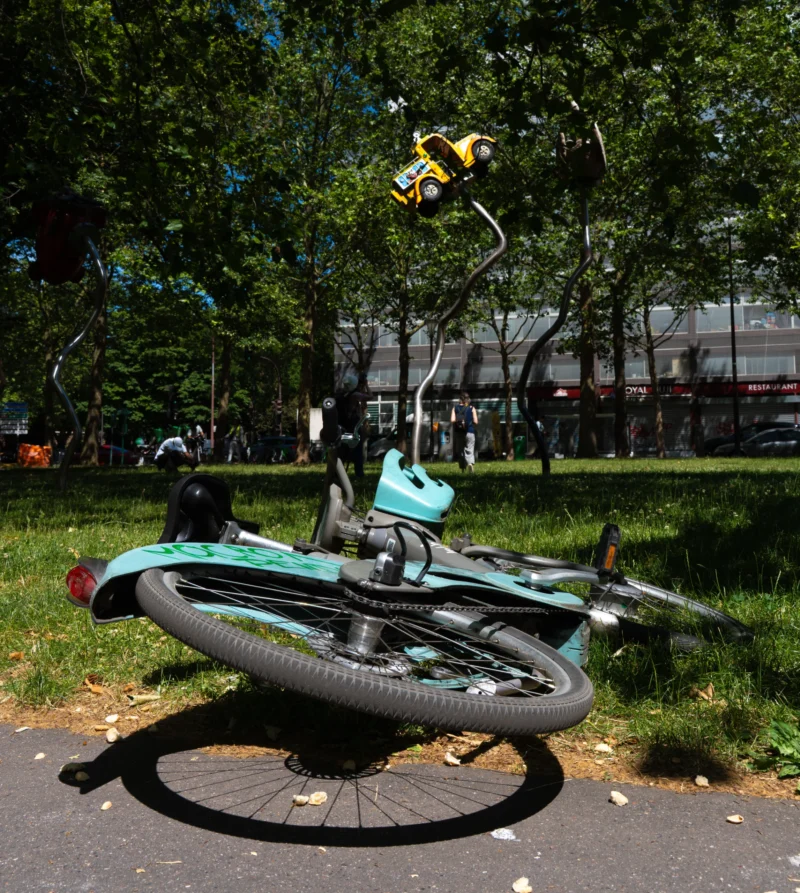 Marc L. · 14/06/2026 · 4672 x 5217 Cette photographie met en scène un vélo turquoise renversé au premier plan, sujet principal. La composition joue sur le contraste entre le vélo et l’arrière-plan flou, avec un cadrage serré sur le vélo et ses accessoires (panneaux solaires, sacs).La lumière naturelle, probablement en journée, éclaire la scène sans ombres marquées. Les couleurs dominantes sont les teintes vives du vélo (turquoise, noir) et les verts des arbres en arrière-plan.Le style est documentaire et spontané, évoquant une ambiance urbaine décontractée et un peu désordonnée.