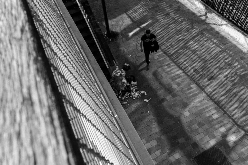 Marc L. · 14/06/2026 · 7008 x 4672 Cette photographie en noir et blanc capture une scène urbaine vue d’en haut. Le sujet principal est un homme marchant avec un chien en laisse, encadré par des murs de briques et une grille métallique. La composition est symétrique, avec des lignes droites et une perspective descendante.Le cadrage serré isole les personnages, créant une ambiance intimiste. La lumière, douce et contrastée, accentue les ombres et les textures. Les couleurs dominantes, bien que monochromes, jouent sur les nuances de gris et de noir, renforçant le côté dramatique et mélancolique. Le style est documentaire et épuré, évoquant une atmosphère solitaire et contemplative.