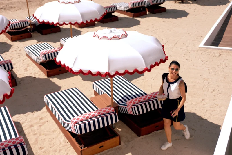 Fabienne · 8468 x 5646 Cette photographie montre une femme comme sujet principal, marchant sur une plage. La composition inclut plusieurs transats noirs et blancs sous des parasols à rayures rouges et blanches. Le cadrage est serré sur la femme, avec un arrière-plan légèrement flou.La lumière semble naturelle et ensoleillée, créant des ombres douces. Les couleurs dominantes sont les tons blancs, noirs et rouges, apportant une ambiance estivale et élégante. Le style est décontracté mais soigné, évoquant une scène de détente en bord de mer. L’ambiance est relaxante et joyeuse.