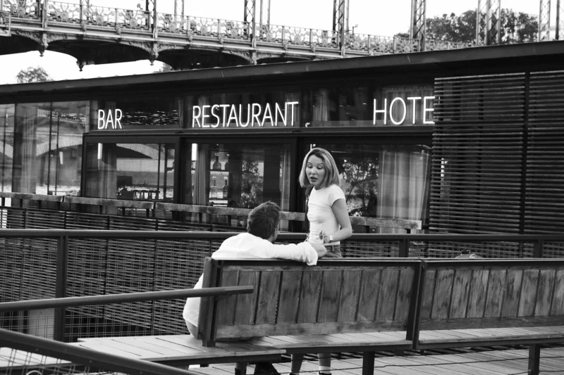Dominique · 23/05/2026 · 2000 x 1333 Cette photographie en noir et blanc met en scène un couple assis sur un banc devant un établissement affichant "Bar", "Restaurant" et "Hotel". Le sujet principal est le couple, dont la femme porte une robe légère et un chapeau, tandis que l’homme semble détendu.La composition est équilibrée, avec le banc et les enseignes en arrière-plan. Le cadrage est serré sur le couple, mais inclut aussi des éléments architecturaux et urbains. La lumière est douce, probablement naturelle, créant des contrastes modérés. Les tons dominants sont les gris et noirs, typiques du noir et blanc, renforçant une ambiance rétro et intemporelle. L’atmosphère évoque la détente et la nostalgie.