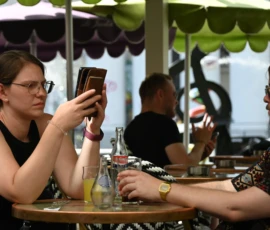 Pierre · 30/05/2026 · 5172 x 3444 Cette photographie montre trois personnes dans un café en terrasse. Le sujet principal semble être la jeune femme à gauche, absorbée par son téléphone. La composition met en avant un cadrage serré sur les visages et les mains, avec une légère profondeur de champ.La lumière est douce et naturelle, probablement en soirée. Les couleurs dominantes sont des tons neutres (noirs, beiges) avec des touches de vert (feuilles) et de bleu (boissons). L’ambiance est calme et intimiste, évoquant une détente urbaine. Le style est réaliste et documentaire.