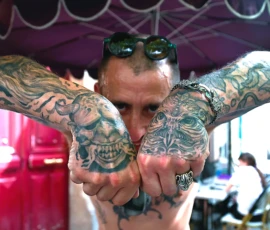 Pierre · 30/05/2026 · 5129 x 3413 Cette photographie met en scène un homme au **sujet principal** très marqué par des **tatouages** couvrant ses bras, notamment des motifs crâniens et symboliques. La **composition** est centrée sur ses bras levés, encadrés par un auvent coloré.Le **cadrage** est serré, mettant en valeur les détails des tatouages. La **lumière** semble naturelle, bien que légèrement contrastée, accentuant les textures de la peau et des motifs.Les **couleurs dominantes** sont les teintes sombres des tatouages, contrastant avec les tons chauds et vifs de l’arrière-plan (rouge, violet). Le **style** est réaliste et documentaire, créant une **ambiance** intense et mystérieuse, évoquant une culture alternative et artistique.