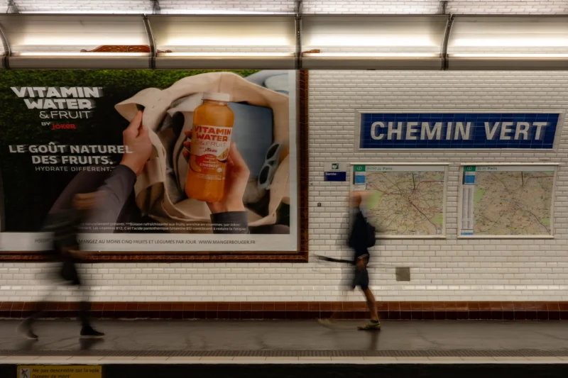 Jean-Marc · 11/06/2026 · 4367 x 2911 Cette photographie montre deux personnes floues en mouvement dans un métro. Le sujet principal est une affiche publicitaire pour "Vitamin Water", mettant en scène un fruit et un texte en français ("Le goût naturel des fruits"). La composition inclut aussi un panneau indiquant "Chemin Vert" avec un plan.Le cadrage est serré sur l’affiche et les voyageurs. La lumière est naturelle et tamisée, typique d’un environnement souterrain. Les couleurs dominantes sont les tons froids (bleu, gris) avec des touches vives (orange, vert) sur l’affiche. Le style est réaliste et documentaire, créant une ambiance urbaine et dynamique.