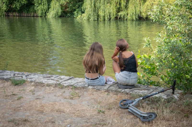 Jean-Marc · 28/06/2026 · 4500 x 3000 Cette photographie montre deux jeunes femmes assises sur une berge herbeuse près d’un plan d’eau. Le sujet principal est leur interaction paisible, entourées de nature.La composition est équilibrée, avec un cadrage serré sur elles et un arrière-plan flou mettant en valeur le lac et les arbres. La lumière est douce et naturelle, probablement en journée, créant des ombres légères. Les couleurs dominantes sont les verts (végétation) et les bleus (eau), avec des touches de beige et de gris.Le style est réaliste et spontané, évoquant une ambiance calme et intime, presque poétique.
