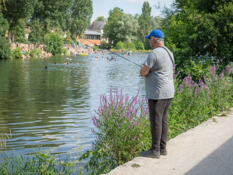 Jean-Marc · 28/06/2026 · 4385 x 2923 Un homme pêche tranquillement au bord d'un plan d'eau entouré de végétation, avec des baigneurs en arrière-plan sous un ciel
