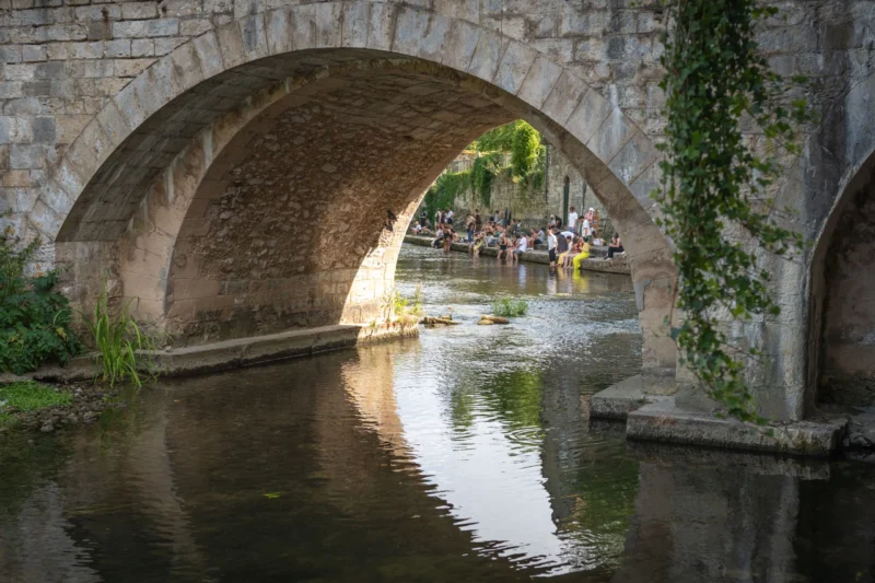 Jean-Marc · 28/06/2026 · 5105 x 3403 Cette photographie met en scène un **pont en pierre à deux arches** enjambant un canal. Le **sujet principal** est l’architecture du pont, reflétée dans l’eau calme.La **composition** joue sur les lignes courbes des arches et la symétrie. Le **cadrage** serré isole le pont, avec une perspective légèrement basse.La **lumière** est douce et naturelle, créant des ombres douces et des reflets dorés. Les **couleurs dominantes** sont les tons gris-beige de la pierre, les verts des végétaux et les reflets dorés de l’eau.Le style est **réaliste et contemplatif**, évoquant une **ambiance paisible et historique**.
