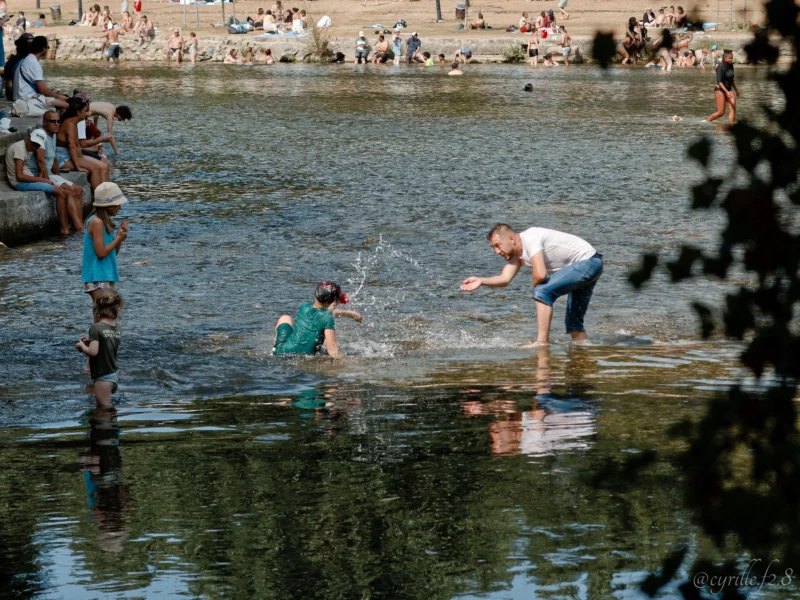 Cyrille · 28/06/2026 · 2026 x 1519 Cette photographie capture une scène de baignade en rivière. Le sujet principal est un groupe de personnes, dont trois hommes en premier plan, partiellement immergés dans l’eau.La composition montre une foule en arrière-plan, suggérant une ambiance conviviale et animée. Le cadrage est serré sur les baigneurs, avec des branches en bas à droite encadrant la scène.La lumière semble naturelle, probablement en journée, avec des ombres douces. Les couleurs dominantes sont les teintes bleutées de l’eau, les tons verts des végétaux et les couleurs vives des vêtements des baigneurs.Le style est documentaire, reflétant une ambiance spontanée et estivale.