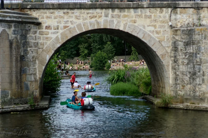 Cyrille · 28/06/2026 · 2026 x 1347 Cette photographie montre un groupe de personnes en kayaks sous un pont en pierre. Le **sujet principal** est l’activité de kayak sur une rivière paisible.La **composition** est dynamique, avec un cadrage en contre-plongée sous l’arche du pont, créant une perspective immersive. La **lumière** naturelle, douce et légèrement tamisée, donne une ambiance sereine. Les **couleurs dominantes** sont les verts des berges et les teintes vives des kayaks, contrastant avec le gris de la pierre.Le **style** est documentaire et naturel, évoquant une **ambiance** détendue et conviviale, mêlant aventure et tranquillité.