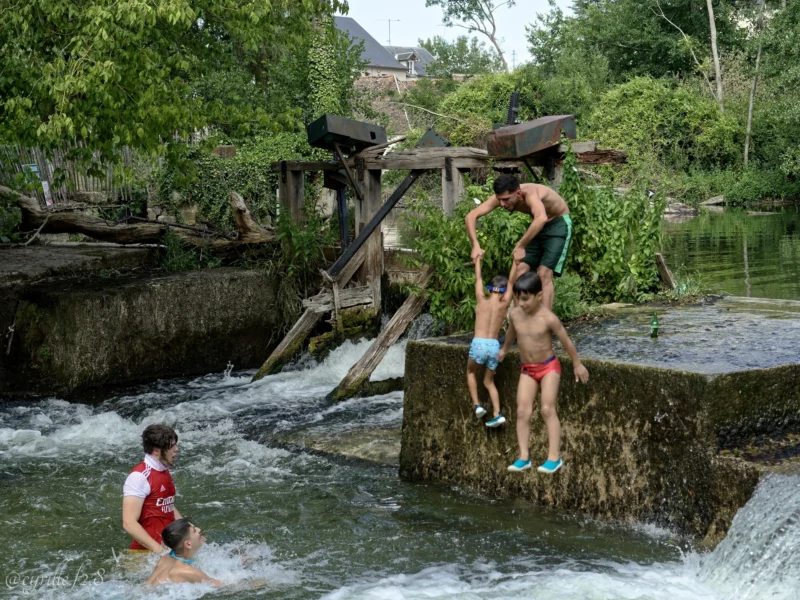 Cyrille · 28/06/2026 · 2026 x 1520 Cette photographie montre des enfants comme sujets principaux, jouant près d’un petit cours d’eau turbulent. La composition met en scène un pont de bois en arrière-plan, avec des enfants grimpant et s’amusant sur les structures environnantes.Le cadrage est dynamique, capturant à la fois l’action et l’environnement naturel. La lumière semble naturelle, probablement en journée, avec des ombres douces. Les couleurs dominantes sont les verts de la végétation, les tons gris-beige des pierres et du bois, et les couleurs vives des vêtements des enfants.Le style est documentaire et spontané, évoquant une ambiance joyeuse et insouciante.