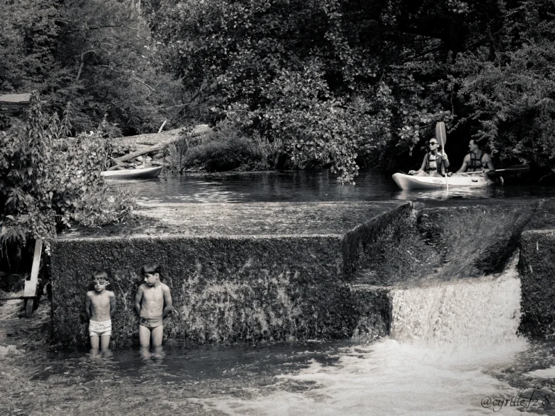 Cyrille · 28/06/2026 · 2026 x 1519 Cette photographie en noir et blanc montre deux enfants comme sujets principaux, debout près d’un petit barrage ou d’une cascade artificielle. La composition met en valeur un paysage naturel avec des arbres et des rochers.Le cadrage inclut une vue large de l’environnement, capturant aussi un bateau à l’arrière-plan. La lumière semble douce et naturelle, typique d’un jour ensoleillé. Les tons dominants sont les gris et noirs, accentuant les contrastes.Le style est documentaire et nostalgique, évoquant une ambiance paisible et simple, presque intemporelle.