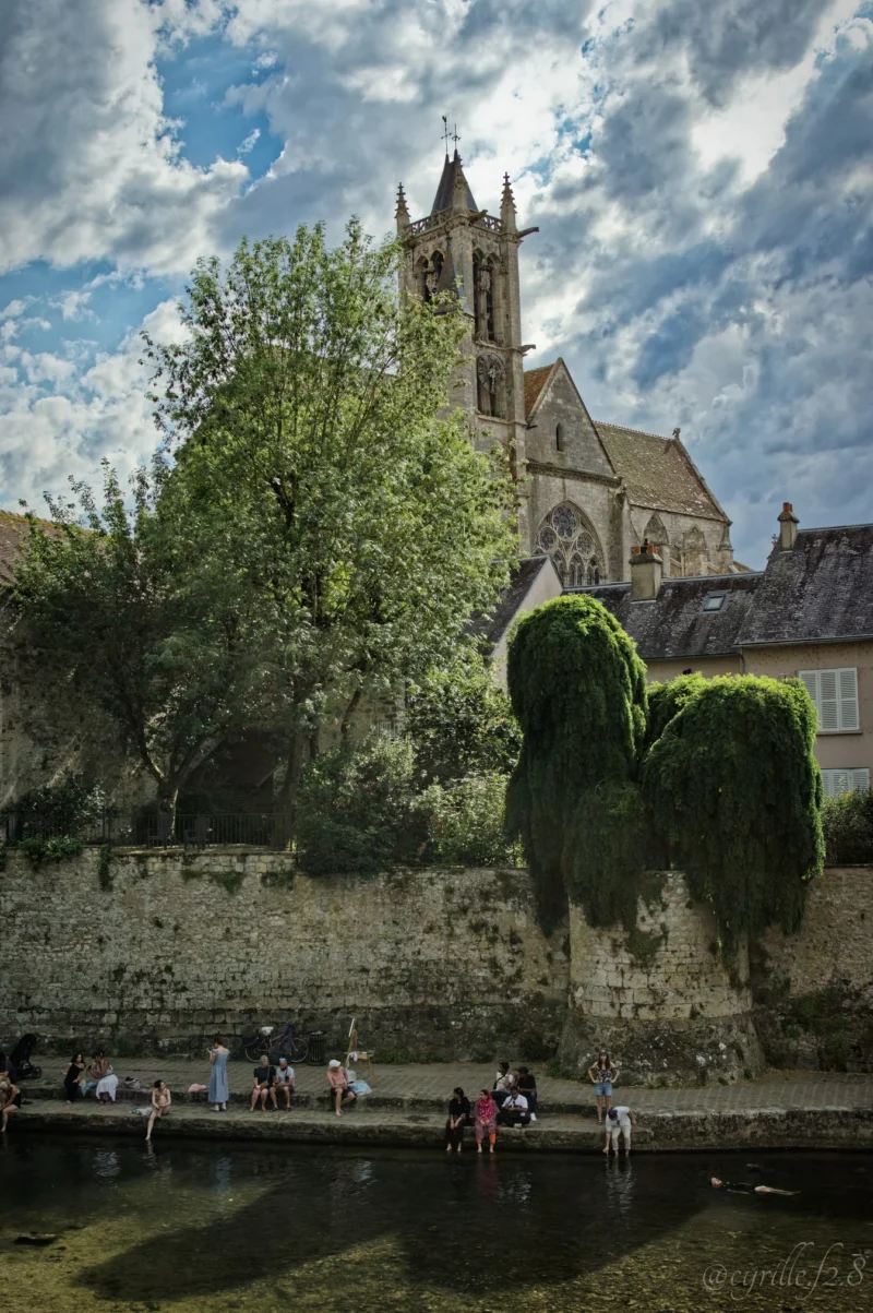Cyrille · 28/06/2026 · 1347 x 2026 Cette photographie met en scène un **pont enjambant un canal**, avec une **statue équestre** comme sujet principal, partiellement cachée par des arbres. La composition joue sur les lignes horizontales du canal et verticales de l’architecture.Le cadrage est serré sur la statue et le pont, intégrant une église gothique en arrière-plan. La lumière, naturelle et douce, crée des contrastes entre ombres et clartés. Les **couleurs dominantes** sont les verts des arbres, les tons gris-beige des pierres et le bleu du ciel.Le style est **réaliste et photographique**, avec une ambiance paisible et légèrement nostalgique.
