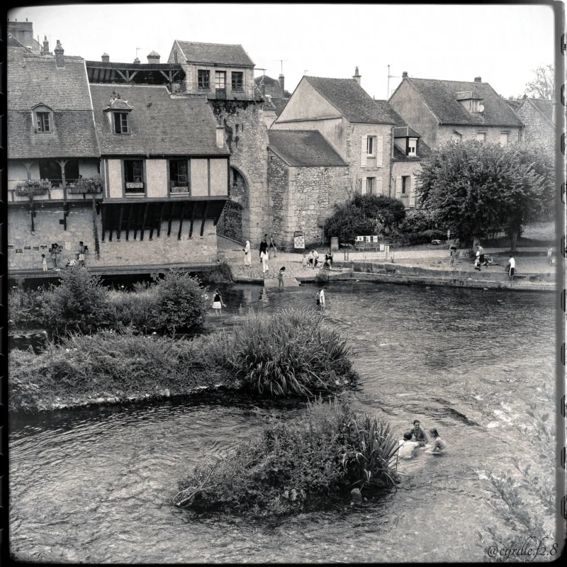 Cyrille · 28/06/2026 · 2026 x 2026 Cette photographie en noir et blanc capture un village européen pittoresque. Le sujet principal est un ensemble de maisons anciennes en bordure d’un ruisseau peu profond. La composition met en valeur les bâtiments aux toits d’ardoise et les fenêtres alignées, encadrés par une végétation riveraine.Le cadrage inclut une vue large sur les habitations et le cours d’eau, avec des personnes en arrière-plan. La lumière semble douce et naturelle, typique d’un jour nuageux. Les tons dominants sont les gris et noirs, accentuant les textures des pierres et des arbres.Le style est documentaire, évoquant une ambiance paisible et intemporelle, presque nostalgique.