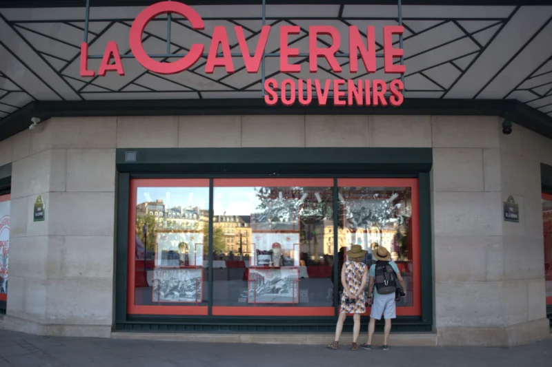 Fabienne · 20/06/2026 · 4895 x 3263 Cette photographie montre une boutique nommée **« La Caverne Souvenirs »**. Le sujet principal est l’entrée du magasin, avec son enseigne rouge et ses vitrines exposant des souvenirs.La composition met en valeur la façade claire et les deux personnes devant la vitrine. Le cadrage est serré sur l’entrée, avec une perspective légèrement basse. La lumière est naturelle et douce, éclairant bien la scène. Les couleurs dominantes sont le rouge (enseigne), le beige (bâtiment) et des tons neutres.L’ambiance est urbaine et touristique, évoquant un lieu dédié aux achats de souvenirs. Le style est réaliste et documentaire.