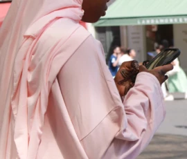 Sophie · 1175 x 1278 Cette photographie met en scène une personne vêtue d’une longue robe et d’un voile roses, observant son téléphone portable. Le sujet principal est flou, ce qui attire l’attention sur ses gestes et son attitude.Le cadrage est serré, centré sur elle, avec un arrière-plan légèrement flou montrant des bâtiments et des passants. La lumière est naturelle, douce et diffuse, créant une ambiance lumineuse et apaisante. Les couleurs dominantes sont les tons pastel roses, contrastant avec les teintes plus neutres de l’arrière-plan.Le style est réaliste et documentaire, capturant un moment du quotidien. L’ambiance est calme et intime, malgré le contexte urbain.