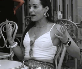 Sophie · 1014 x 1062 Cette photographie met en scène une femme comme sujet principal, assise à une table, souriante et détendue. La composition montre un cadrage serré sur elle, avec des éléments d’arrière-plan flous (autres personnes, objets).La lumière semble naturelle et douce, créant des contrastes modérés. Les couleurs dominantes sont en noir et blanc, ce qui accentue les textures et les jeux de lumière.Le style est informel et spontané, évoquant une ambiance conviviale et décontractée, probablement lors d’un repas ou d’une soirée entre amis.
