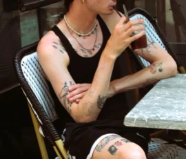 Sophie · 918 x 1379 Cette photographie met en scène un jeune homme assis, qui semble être le sujet principal. Il porte des vêtements minimalistes (soutien-gorge noir et short) et arbore plusieurs tatouages.La composition est simple, avec un cadrage serré centré sur lui, assis sur une chaise en bois. L’arrière-plan flou suggère un café ou un bar. La lumière est douce et naturelle, créant une ambiance tamisée. Les couleurs dominantes sont les tons chauds et neutres, avec des contrastes entre le noir de ses vêtements et ses tatouages.Le style est documentaire et spontané, évoquant une ambiance décontractée et intime.