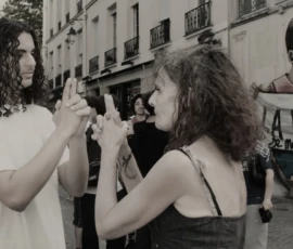 Sophie · 1799 x 1156 Cette photographie en noir et blanc montre deux femmes comme sujets principaux, engagées dans une conversation animée. L’une tient un téléphone, tandis que l’autre semble écouter attentivement.La composition est dynamique, avec un cadrage serré sur les visages et les gestes expressifs. L’arrière-plan, légèrement flou, révèle un bâtiment et une affiche avec le mot "PAIX", suggérant un contexte urbain et militant.La lumière est naturelle, douce, mettant en valeur les traits des femmes. Le style est documentaire, avec une ambiance authentique et spontanée, renforcée par le monochrome.
