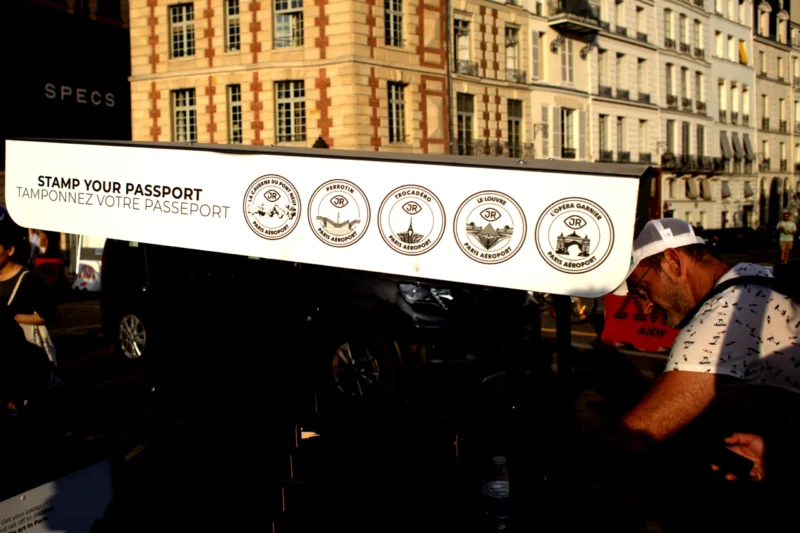 Fabienne · 28/06/2026 · 4593 x 3062 Cette photographie montre un groupe de cyclistes participant à une manifestation. Le sujet principal est une banderole avec le slogan *« Stamp Your Passport »* et des logos d'événements cyclistes. La composition est dynamique, avec les vélos en premier plan et un immeuble en arrière-plan.Le cadrage est serré sur les cyclistes et la banderole. La lumière semble naturelle, avec des ombres douces. Les couleurs dominantes sont les tons neutres (beige, gris) de l'immeuble et les couleurs vives des logos et vêtements des cyclistes. L’ambiance est militante et sportive, évoquant une mobilisation collective.