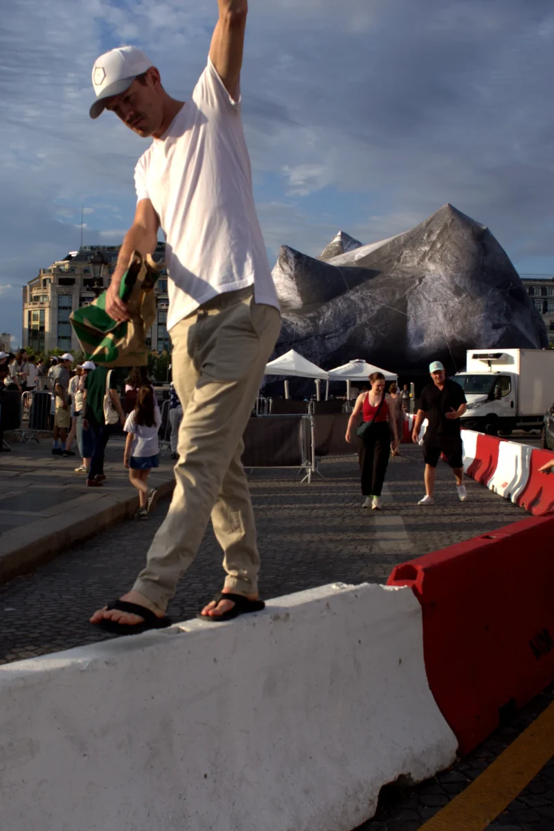 Fabienne · 28/06/2026 · 2359 x 3539 Cette photographie met en scène un homme en mouvement, levant un objet lourd (probablement un poisson géant). La composition est dynamique, avec un cadrage serré sur le sujet principal, isolé sur un premier plan.La lumière semble naturelle, avec des contrastes marqués. Les couleurs dominantes sont les tons clairs (blanc du t-shirt, gris du sol) et des touches de rouge et bleu en arrière-plan. L’ambiance est festive et sportive, évoquant une compétition ou une démonstration de force. Le style est documentaire et spontané.