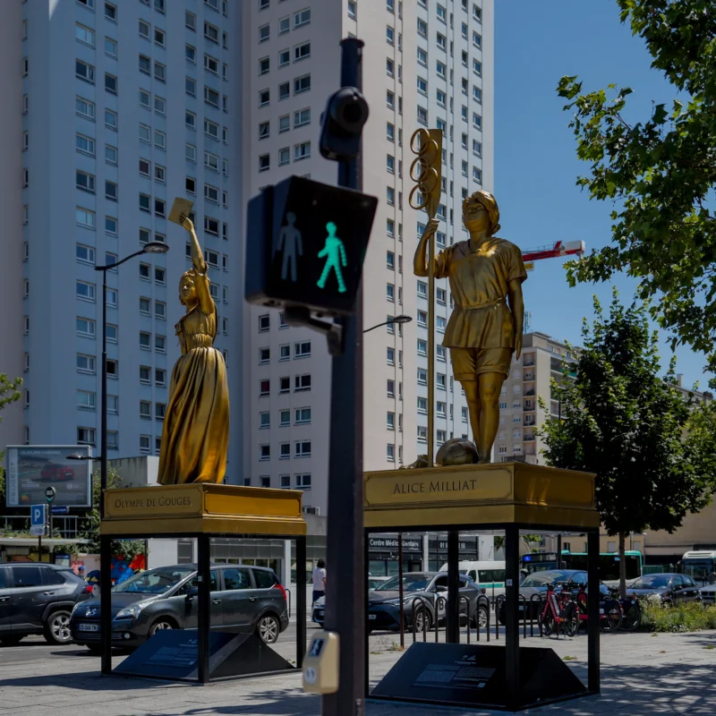 Frédéric · 14/06/2026 · 1680 x 1680 Cette photographie met en scène deux statues dorées représentant des personnages en posture dynamique, placées sur des socles noirs près d’un passage piéton éclairé. Le sujet principal est ces sculptures stylisées, évoquant le mouvement et l’énergie.La composition est symétrique, avec un cadrage serré sur les statues et un arrière-plan urbain (bâtiments, voitures, arbres). La lumière naturelle, vive et contrastée, accentue les reflets dorés et les ombres nettes. Les couleurs dominantes sont le doré des statues, le gris des bâtiments et le vert des arbres. L’ambiance est moderne et artistique, mêlant urbanité et créativité.