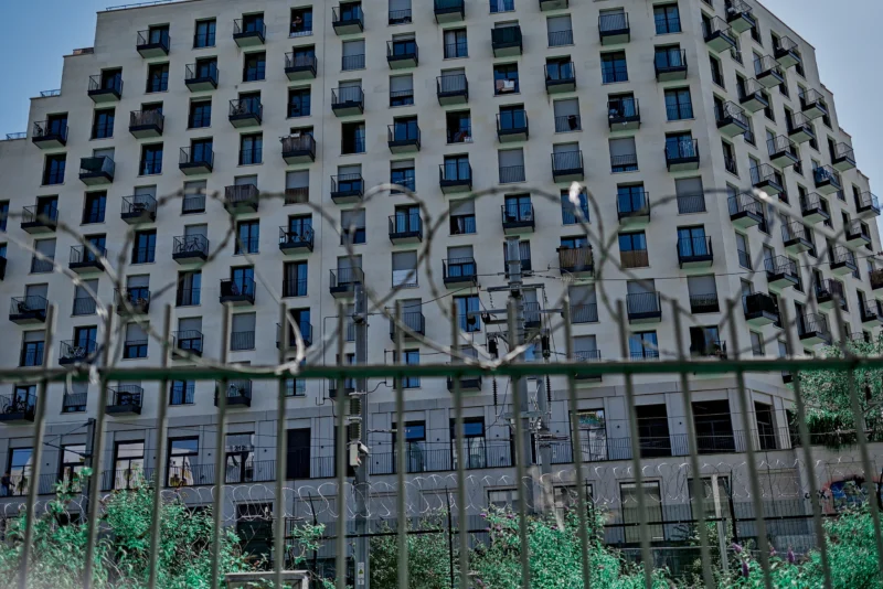 Frédéric · 14/06/2026 · 2517 x 1680 Cette photographie montre un immeuble moderne de grande hauteur, sujet principal. La composition met en valeur sa façade rectiligne et ses nombreux balcons. Le cadrage est serré, capturant presque toute la structure.La lumière semble tamisée, créant des ombres douces. Les couleurs dominantes sont les tons gris-beige de l'immeuble, contrastant avec le vert des arbres en bas. Le style est réaliste et urbain, évoquant une ambiance contemporaine et légèrement mélancolique, renforcée par les jeux de reflets et la lumière diffuse.