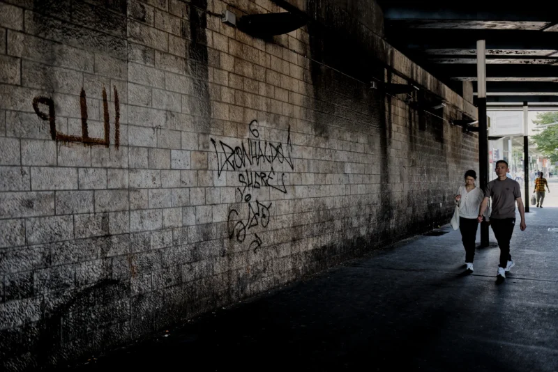 Frédéric · 14/06/2026 · 2517 x 1680 Cette photographie montre deux personnes marchant sous un passage couvert en béton. Le sujet principal est leur silhouette, capturée en contre-jour.La composition est axée sur une perspective longitudinale, avec un cadrage serré des murs couverts de graffitis, dont certains en arabe. La lumière, naturelle et tamisée, crée des ombres marquées, renforçant l’ambiance. Les couleurs dominantes sont les tons gris-beige du béton et les touches sombres des graffitis.Le style est réaliste et documentaire, évoquant une atmosphère urbaine et légèrement mélancolique, voire introspective.