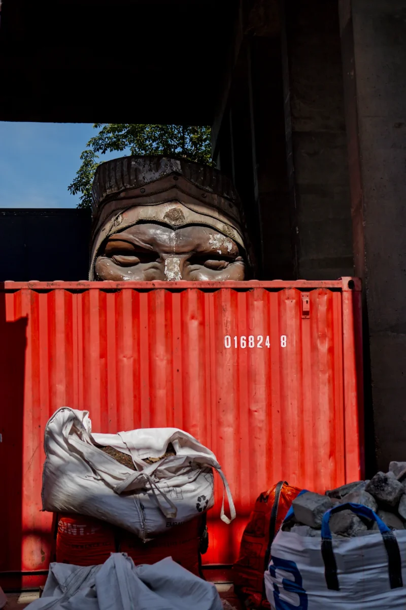 Frédéric · 14/06/2026 · 1120 x 1680 Cette photographie met en scène une tête sculptée, probablement bouddhiste, partiellement cachée derrière un conteneur rouge. Le sujet principal est cette tête, aux traits stylisés et apaisants.La composition joue sur le contraste entre l’art sacré et l’industrie, avec le conteneur métallique en arrière-plan. Le cadrage est serré, centré sur la tête et le conteneur. La lumière naturelle, douce et légèrement contrastée, accentue les détails du visage et les ombres.Les couleurs dominantes sont le rouge vif du conteneur et les tons neutres de la sculpture. Le style est documentaire, mêlant sacré et profane, créant une ambiance à la fois mystérieuse et contemplative.