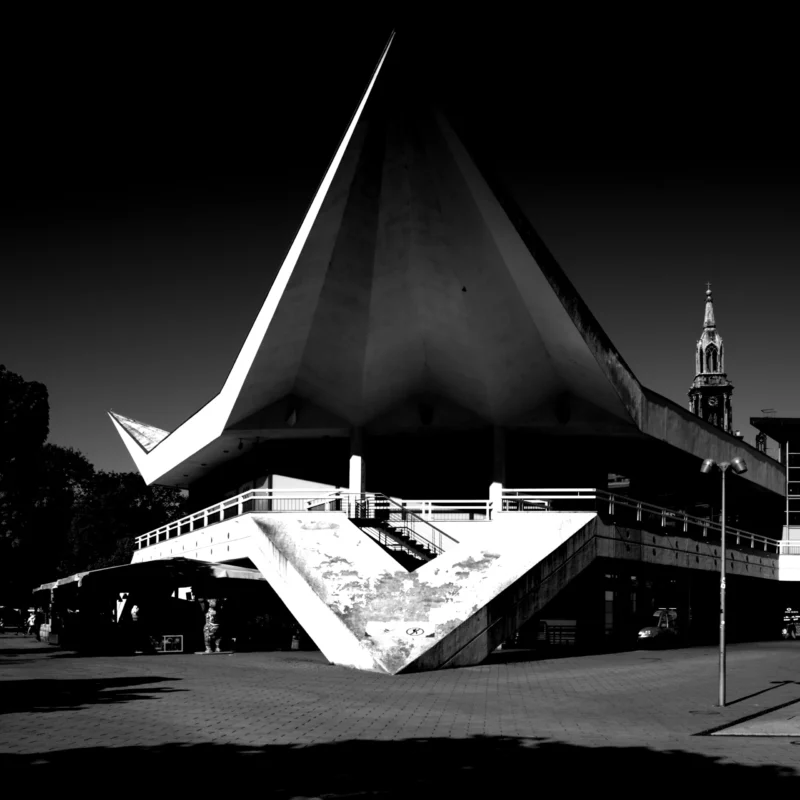 Fabienne · 01/05/2026 · 3636 x 3636 Cette photographie met en scène un bâtiment moderne au design architectural futuriste, marqué par une structure en forme de voile ou de coque penchée. La composition est centrée sur ce sujet principal, avec un cadrage serré qui accentue ses lignes dynamiques.La lumière, contrastée, crée des ombres marquées, renforçant le côté dramatique. En noir et blanc, les tons dominants varient du blanc pur aux noirs profonds, soulignant les textures et les formes architecturales. Le style est épuré et contemporain, évoquant une ambiance à la fois mystérieuse et innovante, typique des architectures visionnaires.