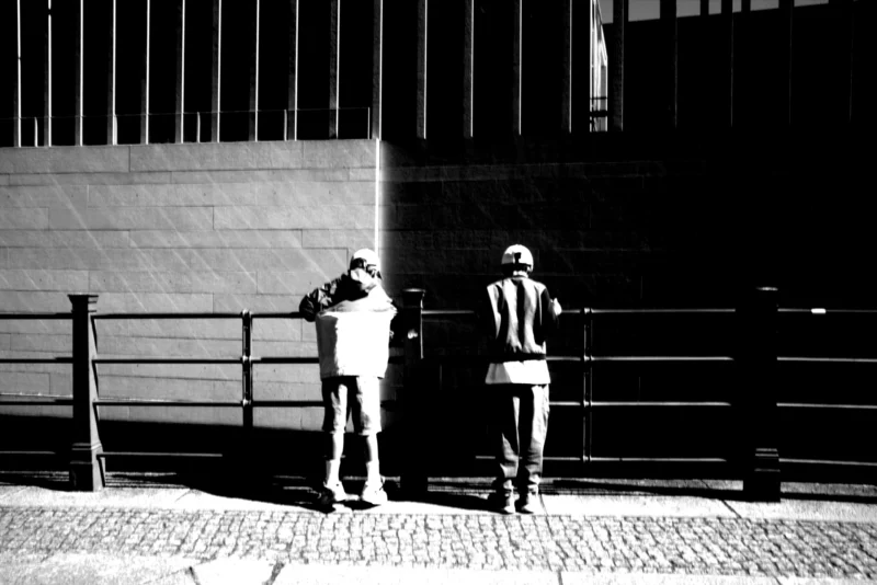 Fabienne · 01/05/2026 · 5496 x 3670 Cette photographie en noir et blanc met en scène deux hommes comme sujets principaux, l’un prenant une photo de l’autre. La composition est épurée, avec un cadrage serré sur les personnages, encadrés par une rambarde métallique. La lumière est douce mais contrastée, créant des ombres marquées. Les tons dominants sont les gris et noirs, renforçant l’aspect dramatique.Le style est documentaire et introspectif, évoquant une ambiance contemplative et presque mélancolique. L’image suggère une réflexion sur la pose et le regard, dans un cadre urbain anonyme.