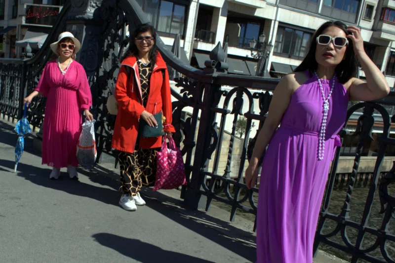 Fabienne · 01/05/2026 · 3662 x 2442 Cette photographie met en scène trois personnes en mouvement près d’un pont. Le sujet principal semble être la femme en robe violette, au premier plan, avec des lunettes et un sourire. La composition est dynamique, avec un cadrage serré sur les visages et les tenues colorées.La lumière est naturelle et vive, suggérant un jour ensoleillé. Les couleurs dominantes sont vives : violet, orange et des tons chauds. Le style est spontané et streetwear, évoquant une ambiance festive et décontractée, presque artistique. L’énergie et la joie transparaissent dans cette scène urbaine.