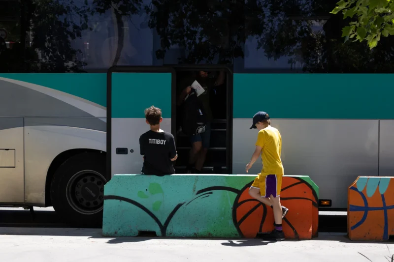 Muriel · 14/06/2026 · 2048 x 1365 Cette photographie met en scène un bus turquoise et des cyclistes en premier plan. Le sujet principal semble être une personne en jaune poussant un vélo, tandis qu’une autre figure, en noir, est partiellement visible près du bus. La composition joue sur les contrastes entre les formes géométriques des vélos et le bus, avec un cadrage serré sur les roues colorées.La lumière est naturelle et semble provenir d’un côté, créant des ombres marquées. Les couleurs dominantes sont les teintes vives (turquoise, jaune, noir) et les tons neutres du sol. Le style est documentaire et urbain, évoquant une ambiance dynamique et moderne dans un espace public.
