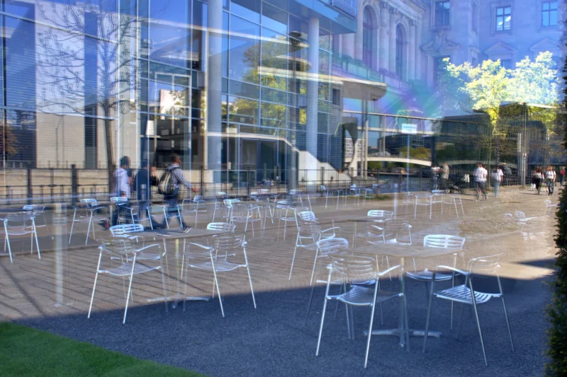 Fabienne · 01/05/2026 · 4310 x 2874 Cette photographie montre un espace urbain moderne avec des tables et chaises blanches disposées devant une façade vitrée reflétante. Le sujet principal est l’architecture et l’aménagement extérieur, mettant en valeur un lieu de détente ou de restauration.La composition est symétrique, avec des lignes droites et un cadrage serré sur les tables. La lumière est naturelle, douce et diffuse, créant des reflets sur les vitres. Les couleurs dominantes sont les tons gris (bâtiment), blancs (mobilier) et bleus (ciel). Le style est épuré et contemporain, évoquant une ambiance calme et sophistiquée, typique d’un espace public moderne et bien conçu.
