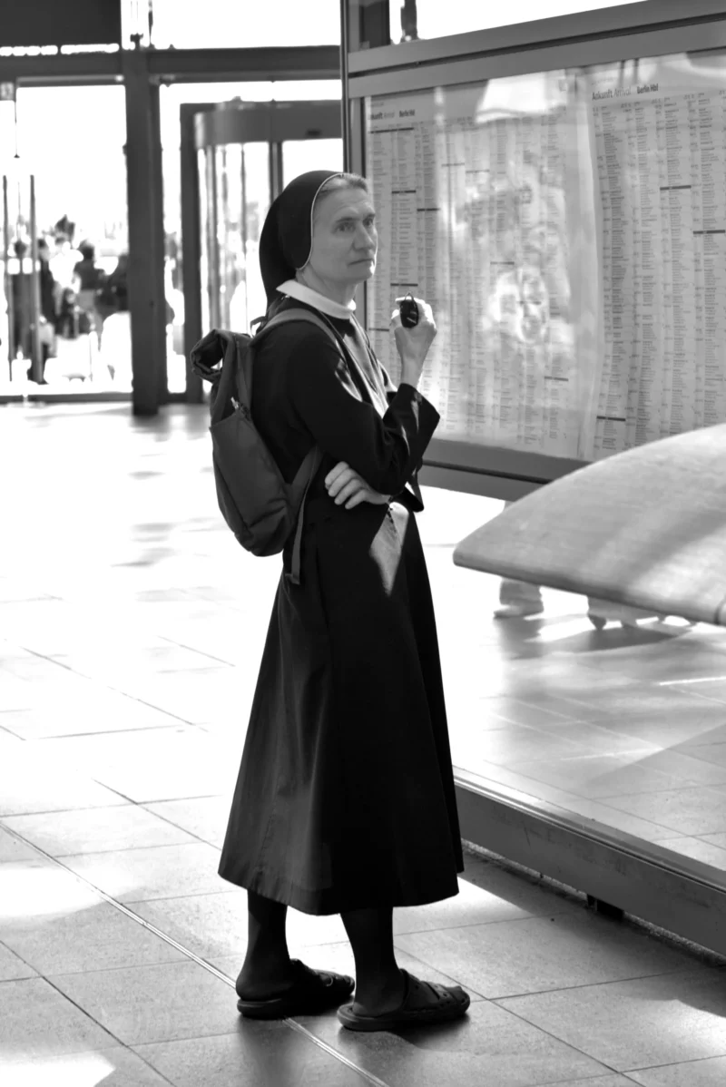 Fabienne · 01/05/2026 · 3670 x 5496 Cette photographie en noir et blanc met en scène une femme vêtue d'une longue robe noire, probablement une religieuse, comme sujet principal. Elle tient un livre ou un objet similaire.La composition est simple et épurée, avec un cadrage serré sur la personne, qui se détache sur un arrière-plan flou et légèrement lumineux. La lumière semble naturelle, douce et diffuse, créant des contrastes modérés.L’ambiance est sobre et contemplative, renforcée par le monochrome. Le style est documentaire, évoquant une scène du quotidien dans un lieu public, probablement une gare ou un hall d’aéroport.