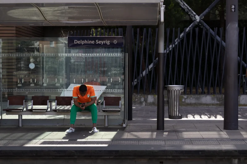 Muriel · 14/06/2026 · 2048 x 1365 Cette photographie montre un homme en combinaison orange et verte, accroupi près d’un passage piéton. Il semble inspecter ou réparer quelque chose.La composition est simple et épurée, avec un cadrage serré sur le sujet. L’arrière-plan inclut une vitrine et un panneau indiquant "Dalpine Sayve". La lumière est naturelle, douce, avec des ombres peu marquées. Les couleurs dominantes sont les tons verts, oranges et gris.Le style est réaliste et documentaire, capturant une scène du quotidien. L’ambiance est calme et professionnelle, évoquant une activité urbaine discrète.