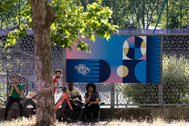 Muriel · 14/06/2026 · 2048 x 1365 Cette photographie montre un groupe de personnes assises sous un arbre, près d’un mur décoré d’un motif géométrique coloré. Le sujet principal est l’interaction sociale dans un cadre urbain détendu.La composition est équilibrée, avec un cadrage serré sur les personnes et le mur coloré en arrière-plan. La lumière naturelle, douce et légèrement filtrée par les feuilles, crée une ambiance chaleureuse. Les couleurs dominantes sont les teintes vives du mur (bleu, rose, jaune) contrastant avec le vert des feuilles et les tons neutres des vêtements.Le style est contemporain et artistique, évoquant une ambiance conviviale et créative.