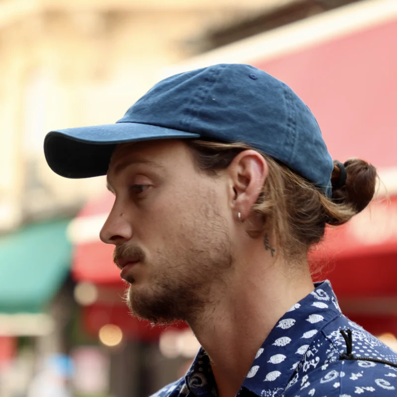 Francoise · 20/06/2026 · 4000 x 4000 Cette photographie met en scène un homme en **portrait serré**, sujet principal au premier plan. Il porte une **casquette bleue** et un **pull à motifs**, avec les cheveux attachés en queue-de-cheval. Le **cadrage** est serré sur son visage et son épaule.La **lumière** semble naturelle, avec des ombres douces. Les **couleurs dominantes** sont les tons bleus (casquette, pull) et rouges (arrière-plan flou). L’ambiance est **urbaine et décontractée**, avec un style **spontané et réaliste**, évoquant une atmosphère de rue animée.