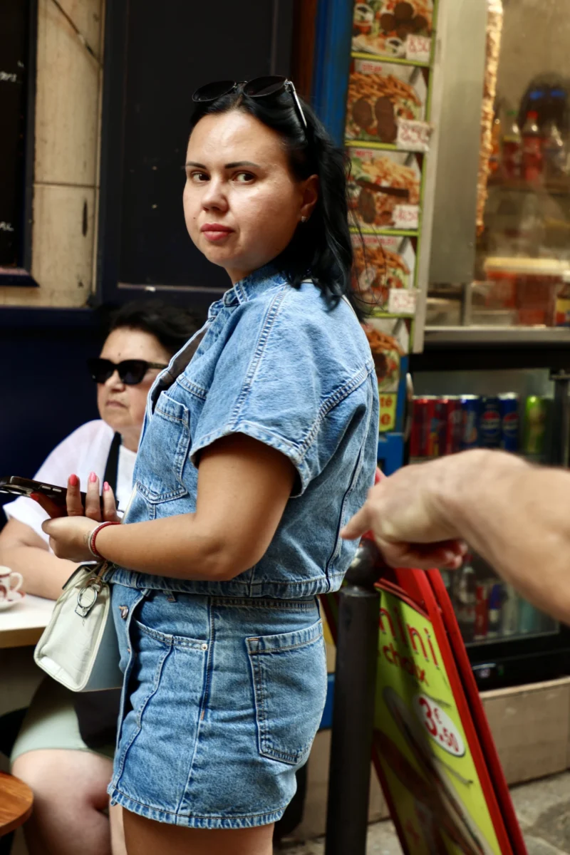 Francoise · 20/06/2026 · 4000 x 6000 Cette photographie montre une jeune femme comme sujet principal, vêtue d’une tenue décontractée (haut bleu et short denim), posant avec un air légèrement détendu. Le cadrage est serré, centré sur elle, avec une autre personne partiellement visible en arrière-plan.La lumière semble naturelle, bien que légèrement tamisée, créant des contrastes doux. Les couleurs dominantes sont les tons bleus et blancs de ses vêtements, avec des touches de couleurs vives en arrière-plan (objets colorés).L’ambiance est informelle et urbaine, évoquant une scène de rue ou un marché. Le style est réaliste et spontané.