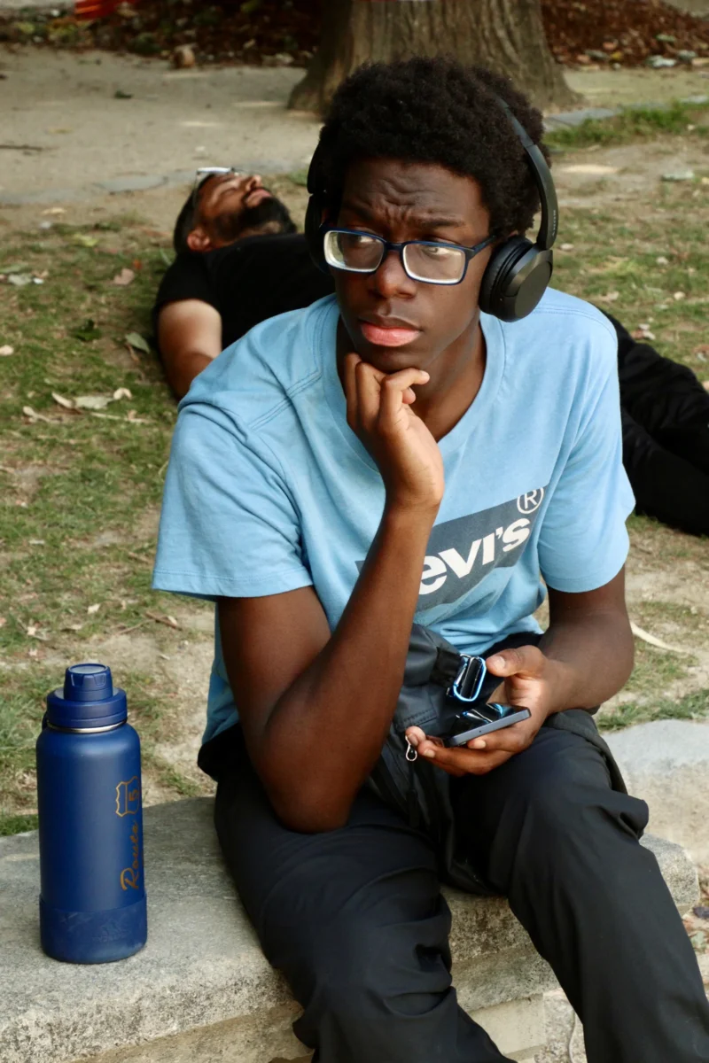 Francoise · 20/06/2026 · 3703 x 5555 Cette photographie montre un jeune homme assis, qui semble réfléchir en tenant son menton. Il porte des lunettes, un t-shirt bleu et des écouteurs.La composition met en avant le sujet principal, légèrement décentré. Le cadrage est serré, capturant aussi un objet bleu (une bouteille) et une partie d’un autre individu en arrière-plan.La lumière est naturelle, douce, avec des ombres peu marquées. Les couleurs dominantes sont les tons bleus et verts, créant une ambiance calme et détendue. Le style est réaliste et spontané, évoquant un moment de pause dans un cadre extérieur.