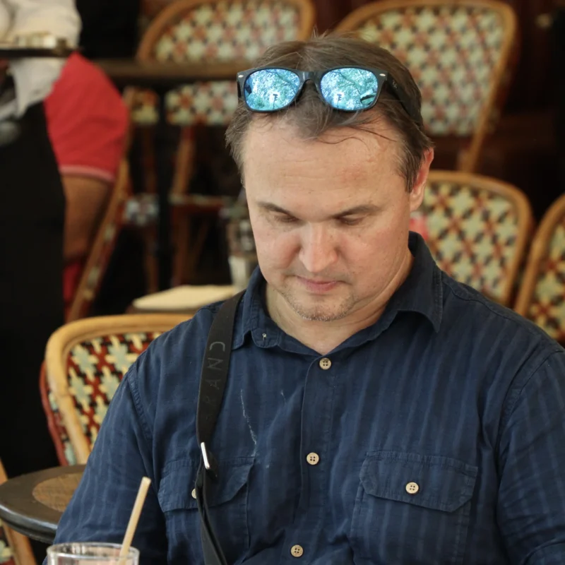 Francoise · 20/06/2026 · 3609 x 3609 Cette photographie montre un homme comme sujet principal, assis à une table en extérieur. Il porte des lunettes de soleil posées sur sa tête et un shirt bleu. Le cadrage est serré, centré sur lui, avec des chaises et des personnes en arrière-plan légèrement floues.La lumière semble naturelle, probablement en journée. Les couleurs dominantes sont les tons bleus du vêtement de l’homme et les teintes chaudes des chaises en osier. L’ambiance est décontractée et informelle, évoquant un cadre convivial, peut-être un café ou un restaurant en plein air.