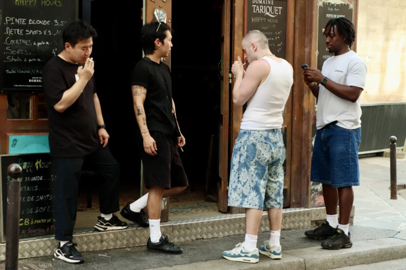 Francoise · 20/06/2026 · 6000 x 4000 Cette photographie capture un groupe de quatre hommes en extérieur devant une enseigne de restaurant. Le sujet principal semble être leur interaction sociale, avec des gestes et postures décontractées.La composition montre une scène urbaine, avec un cadrage serré sur les individus. La lumière est naturelle, probablement en journée, éclairant les visages et créant des ombres douces. Les couleurs dominantes sont neutres et vives : noirs, blancs, gris, et touches de couleurs vives comme le bleu du short et les motifs du t-shirt.Le style est documentaire, réaliste, reflétant une ambiance détendue et spontanée dans un cadre urbain animé.