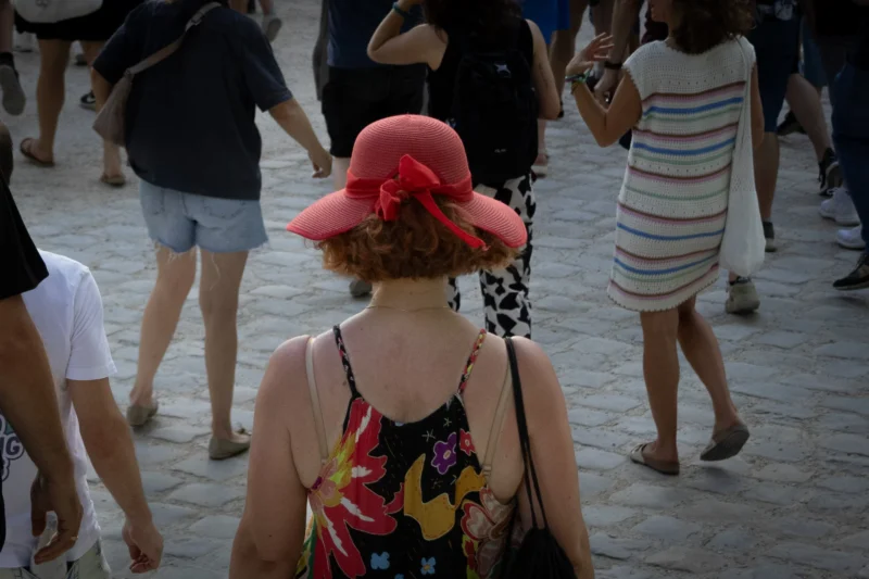 Muriel · 20/06/2026 · 2048 x 1365 Cette photographie montre une femme comme sujet principal, vue de dos, portant un chapeau rouge et un haut coloré. La composition est dynamique, avec plusieurs personnes en mouvement autour d’elle.Le cadrage est serré, centré sur elle mais incluant des éléments périphériques. La lumière semble naturelle, probablement en extérieur, avec des ombres douces. Les couleurs dominantes sont vives : rouge, rose, bleu et blanc.Le style est documentaire et spontané, capturant une scène de vie quotidienne. L’ambiance est estivale et animée, évoquant une promenade en plein air.