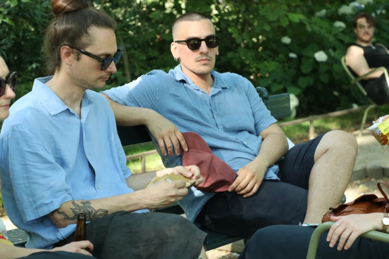 Francoise · 20/06/2026 · 6000 x 4000 Cette photographie montre deux hommes assis sur un banc en extérieur. Le sujet principal est leur interaction détendue, l’un tenant un objet dans les mains.La composition est simple et naturelle, avec un cadrage serré centré sur eux. La lumière semble douce et naturelle, probablement en journée. Les couleurs dominantes sont les tons bleus de leurs vêtements, contrastant avec le vert des arbres en arrière-plan.Le style est réaliste et spontané, évoquant une ambiance décontractée et conviviale, presque intimiste.