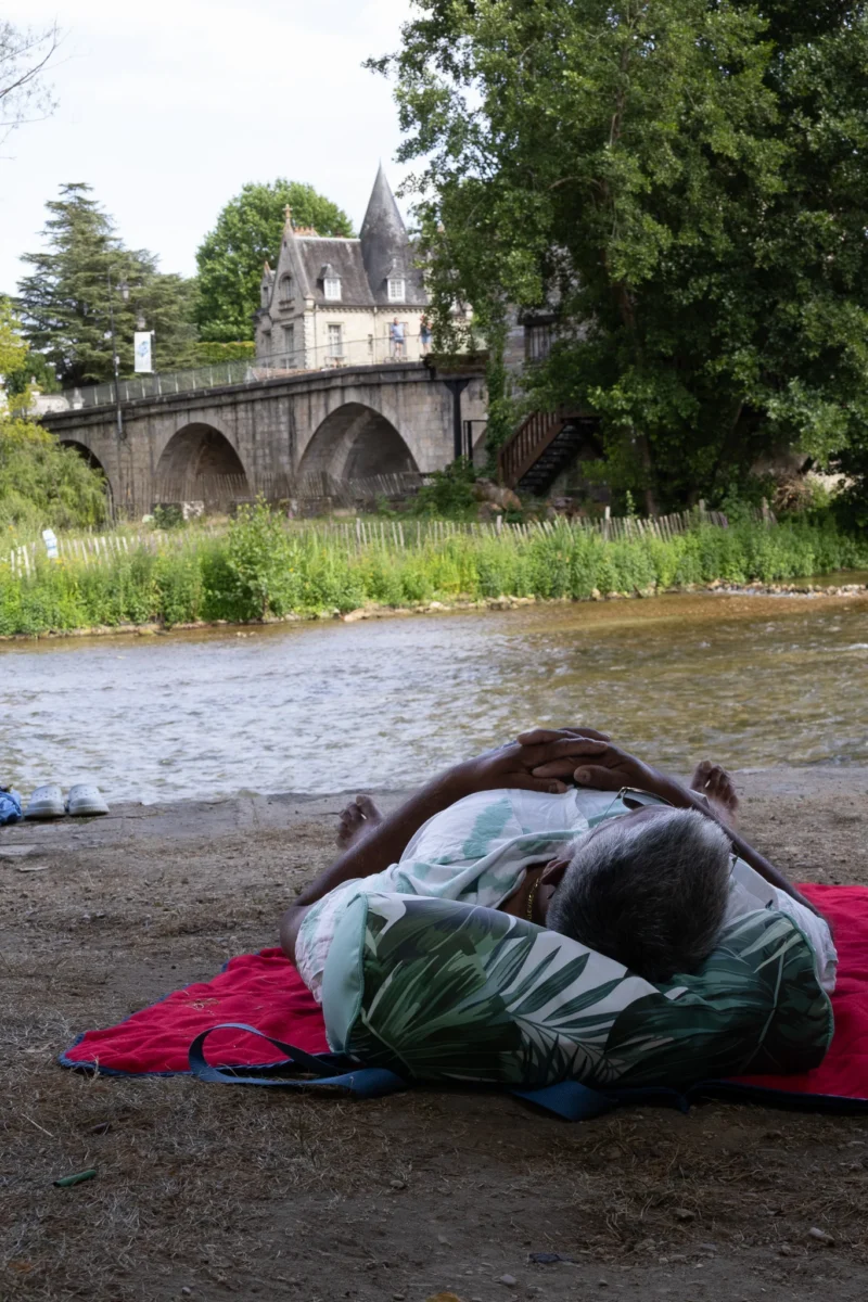 Muriel · 28/06/2026 · 1365 x 2048 Cette photographie montre deux personnes allongées sur une couverture rouge près d’un plan d’eau. Le sujet principal est leur posture détendue, presque endormies.La composition met en valeur l’avant-plan avec les deux individus, tandis que l’arrière-plan révèle un pont en pierre et un château, créant une profondeur. Le cadrage est serré sur les personnes, mais inclut des éléments architecturaux pour le contexte.La lumière semble naturelle, douce et légèrement tamisée, évoquant une ambiance paisible. Les couleurs dominantes sont les verts de la végétation, les tons gris du château et les rouges de la couverture. Le style est réaliste et documentaire, avec une ambiance calme et romantique.