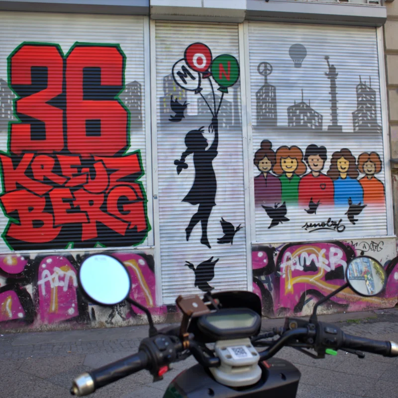 Fabienne · 02/05/2026 · 3670 x 3670 Cette photographie montre un mur tagué en ville. Le sujet principal est un graffiti stylisé avec le numéro "36" et le mot "Kreuzberg", accompagné de dessins colorés : une silhouette en mouvement tenant un ballon et des personnages stylisés.La composition inclut aussi un scooter garé devant. Le cadrage est serré, mettant en valeur les détails des tags. Les couleurs dominantes sont vives : rouge, noir, bleu et vert. Le style est urbain et artistique, reflétant une ambiance dynamique et créative, typique des quartiers alternatifs. La lumière semble naturelle, probablement en journée.