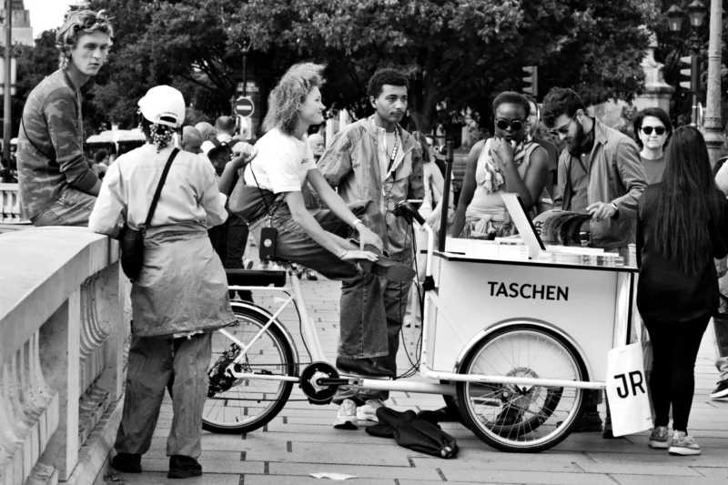 Fabienne · 12/06/2026 · 3651 x 2434 Cette photographie en noir et blanc capture une scène urbaine animée. Le sujet principal est un homme assis sur un vélo attelé à une remorque publicitaire pour la marque "TASCHEN".La composition montre plusieurs personnes autour, certaines observant ou discutant. Le cadrage est serré sur le vélo et la remorque, avec des éléments architecturaux en arrière-plan. La lumière semble naturelle, probablement en journée, créant des contrastes nets. L’ambiance est dynamique et informelle, évoquant une activité commerciale ou un événement public. Le style est documentaire, reflétant une scène authentique de la vie quotidienne.