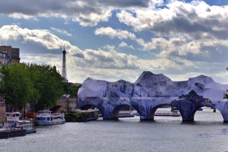 Fabienne · 12/06/2026 · 4401 x 2934 Cette photographie met en scène un pont urbain recouvert d’une installation artistique évoquant des montagnes ou des rochers. Le sujet principal est cette structure transformée, créant un contraste saisissant avec l’architecture environnante.La composition joue sur l’horizontale du fleuve et la verticalité des bâtiments. Le cadrage inclut une partie de la rive gauche avec des arbres et des bateaux, ajoutant de la profondeur. La lumière, douce et naturelle, souligne les contours et les textures. Les couleurs dominantes sont les bleus du ciel et de l’eau, les gris urbains, et les teintes bleutées de l’installation.Le style est à la fois réaliste et artistique, évoquant une ambiance à la fois moderne et poétique.