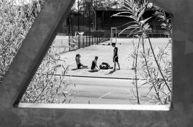 Laurence · 14/06/2026 · 1372 x 900 Cette photographie en noir et blanc montre un groupe de personnes jouant au tennis sur un court extérieur. Le sujet principal est le match en cours, avec des joueurs en action.La composition est dynamique, avec un cadrage légèrement incliné depuis une fenêtre ou une ouverture. La lumière naturelle éclaire la scène, créant des contrastes marqués. Les couleurs dominantes sont les tons de gris, typiques du noir et blanc.Le style est documentaire et spontané, capturant un moment authentique. L’ambiance est sportive et détendue, renforcée par le cadre naturel environnant.