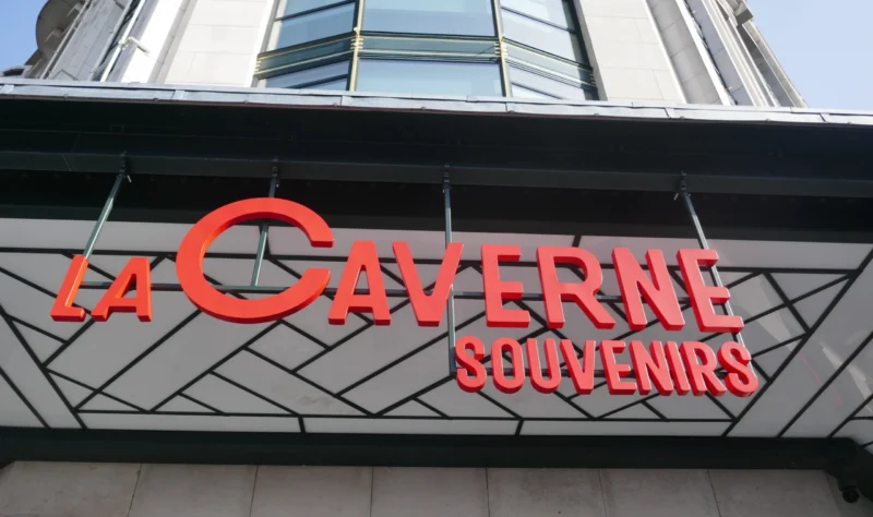Bao · 5472 x 3240 Cette photographie montre principalement l'enseigne d'un magasin nommé **« La Caverne Souvenirs »**. Le sujet principal est le logo stylisé en lettres rouges sur un fond noir et grisé.La composition est épurée, centrée sur l’enseigne. Le cadrage est serré, mettant en valeur les éléments textuels. La lumière semble naturelle, éclairant uniformément la façade. Les couleurs dominantes sont le rouge vif pour le texte et le gris anthracite pour le fond.Le style est moderne et commercial, évoquant une ambiance touristique et accueillante.